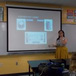 A woman in a yellow skirt presents a slide titled "Experiment Test Chamber" to a classroom, pointing at the slide with a display pointer.