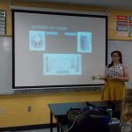 A woman stands in a classroom giving a presentation on an experiment test chamber. She holds a mechanical component and gestures towards a screen displaying diagrams. Students watch from their desks.