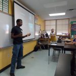 A man stands and speaks in front of a classroom, while a woman and three students sit and listen. A presentation is projected on a screen behind the speaker.