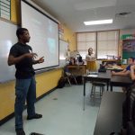 A man is presenting to three students seated at desks in a classroom. A woman stands near the back, observing. Text and images are projected onto a screen behind the presenter.