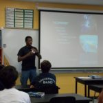 A man stands in front of a classroom, giving a presentation. Behind him is a screen displaying a slide titled "THE EXPERIMENT" with images and text. Several students are seated and watching attentively.