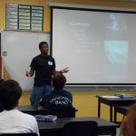 A man is presenting an experiment in a classroom. A projection displays information behind him, and students are seated, facing him. Educational posters are visible on the walls.