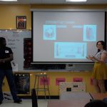 Two presenters stand in a classroom in front of a screen that reads "EXPERIMENT TEST CHAMBER." The person on the right points to a diagram on the screen, while the person on the left observes.