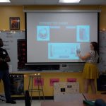 Two people present an experiment test chamber diagram in a classroom. One stands near a stool, the other points at the projection screen displaying detailed images and diagrams.