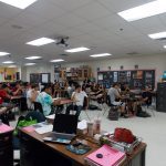 A teacher addresses students seated at desks in a classroom. Various educational materials are visible on the desk in the foreground and posters adorn the walls in the background.