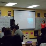 A classroom with students watching a presentation on a screen. Two people are standing at the front, one pointing at the screen that displays a slide titled "The Experiment.