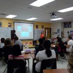 A classroom with students seated at desks while two individuals stand at the front near a projector screen displaying a presentation. Educational posters and charts decorate the walls.