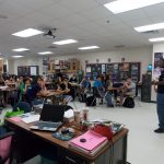 A classroom with students seated at desks, listening to a speaker standing in front. The room has various posters, bookshelves, and a projector hanging from the ceiling.