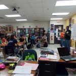 A classroom with students seated at desks while a person at the front gives a presentation. The room has posters on the walls, a projector screen, and various items on the teacher's desk.