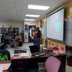 A classroom with students seated at desks, two presenters at the front near a projection screen, and desks filled with papers and supplies. The classroom walls are adorned with posters and whiteboards.