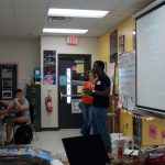 Two people stand in front of a classroom, speaking to students. A presentation is projected on the board. Students sit at desks, listening attentively. Classroom decorations and safety posters are visible.