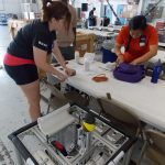 Three people work together on a mechanical project in an indoor workshop, with tools and supplies laid out on a table.