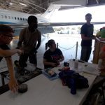 Group of people in a hangar working on a project; one focuses on an item, others discuss around a table covered with equipment and papers; an airplane is partially visible in the background.