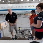 Three individuals work on a project in an aircraft hangar, with a large plane visible in the background. The focal point is a table with equipment in the foreground.