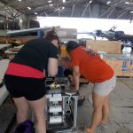 Three adults work on a piece of equipment inside a large hangar, with aircraft and various tools visible around them.