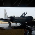 A jet airplane is seen indoors in a hangar with its canopy open. A person is working on a project in the foreground, partially obscured by wooden equipment.