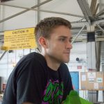 A man is seen indoors with industrial equipment and caution signs in the background. He is wearing a black shirt with green and pink graphic text.