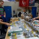 Four people work together on assembling or inspecting a series of scientific or technical equipment on a lab table. Various lab tools and devices are spread across the table.