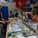 Two people work at a table with multiple scientific instruments and equipment in a laboratory. The man on the left observes while the woman on the right adjusts a device.