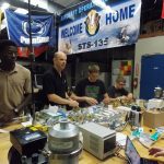 A group of men working on mechanical equipment at a cluttered workstation, with banners reading "WELCOME HOME" and "STS-135" in the background.