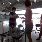 Two women stand in an aircraft hangar, one in workout clothes adjusting straps on her arms, and the other in casual wear assisting her. A piece of equipment with labels is in the foreground.