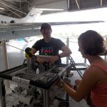 Two individuals work on a piece of equipment inside a hangar, with a partial view of an aircraft in the background. They appear focused and are engaged in a technical task.