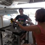 Two people are working on a mechanical device in an aircraft hangar, with a jet situated in the background. One person is wearing a "Theta Tau" shirt.