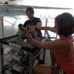 Two individuals work on a mechanical structure in a hangar with an aircraft in the background. One person kneels while adjusting a component, and the other stands behind, holding tools.