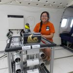 A person in an orange shirt sits inside an aircraft next to scientific equipment labeled "PORT". The equipment is mounted on a metal frame. The aircraft seats are visible in the background.