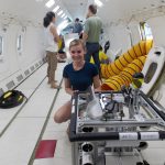 Person kneeling inside an aircraft while working with scientific equipment. Other individuals are standing and conversing in the background.