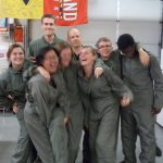 A group of eight people in military-style jumpsuits pose together in a casual manner inside a room with various equipment and flags. Some are smiling and laughing.