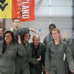 Seven people in gray jumpsuits pose playfully in a group. They are standing in an indoor space with various flags hanging in the background.