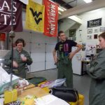 Four people are gathered in a workshop or lab space, wearing jumpsuits. Various university banners hang from the ceiling, including Texas State, Maryland, West Virginia, and Rice University.