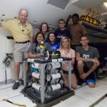 A group of eight people poses around scientific equipment inside an aircraft. Some are kneeling, while others stand. The setting appears to be the interior of a plane, with seats visible in the background.