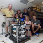 A group of eight individuals, consisting of six young adults and two adults, pose with scientific equipment in the cabin of an aircraft.