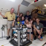 A group of people pose enthusiastically around a robotic device inside an aircraft. There are eight individuals showing various cheerful expressions, and aircraft seats are visible in the background.