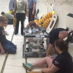 A group of people working on a metallic device inside a confined space with a few seated on the ground and others standing around.