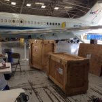 An airplane inside a hangar with wooden crates and equipment scattered around. Tables and chairs are nearby, indicating work in progress.