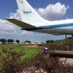 A NASA airplane with the tail number N930NA is parked on a grassy area under a blue sky. There's a small purple stuffed toy placed near the tail of the plane.