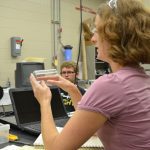 A woman in a lab setting holds a glass container while speaking, with a computer and lab equipment visible. Another person is seated in the background.