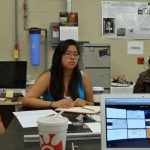 Three people sitting around a table in an office, engaged in discussion. A laptop, documents, and a coffee cup are on the table. Posters and a calendar are visible in the background.