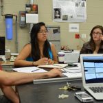 Three people sit at a table with papers, notebooks, and a laptop, engaged in a discussion in a laboratory setting. Various equipment and posters are visible in the background.