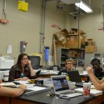 A group of students sits around a table with laptops and notebooks in a lab or classroom, listening to an instructor. The room has lab equipment, boxes, and a mix of industrial and academic settings.