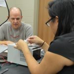 A woman works on an electronic device at a lab bench while a man watches intently.