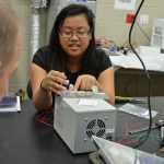 A woman works on an electronic device at a lab bench while a man looks on. Various tools, wires, and equipment are scattered across the desk.