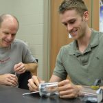 Two men at a table work on assembling a small object. The older man, dressed in a gray t-shirt, watches as the younger man, wearing a green polo, smiles while handling the object.