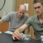 Two men in a laboratory setting are conducting an experiment; one man observes while the other handles a piece of equipment.