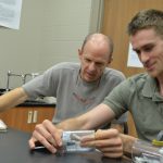 Three people are engaged in a scientific experiment in a lab. One person is holding and examining a small cylindrical object while the other two observe. Laboratory equipment and papers are visible on the desk.