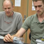 Two men are working on an engineering project at a table. The older man is observing while the younger man is handling a clear cylindrical object. Both are wearing gray shirts.