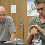 Two men are working at a lab table. One is explaining or demonstrating something with his hands, while the other watches intently. Various scientific equipment and diagrams are in the background.